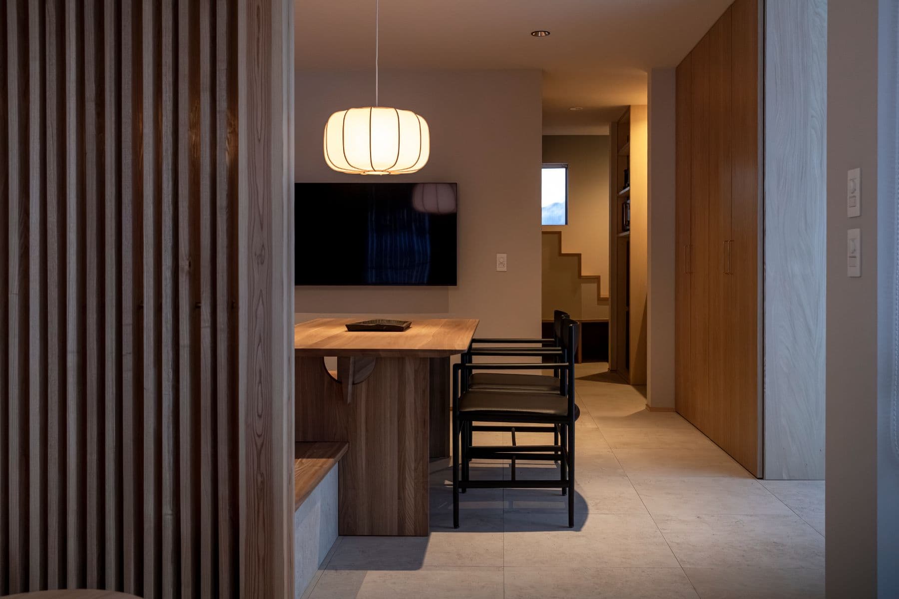 A Japanese-modern dining area with a wooden table and black chairs, with a wood-slat partition and staircase.