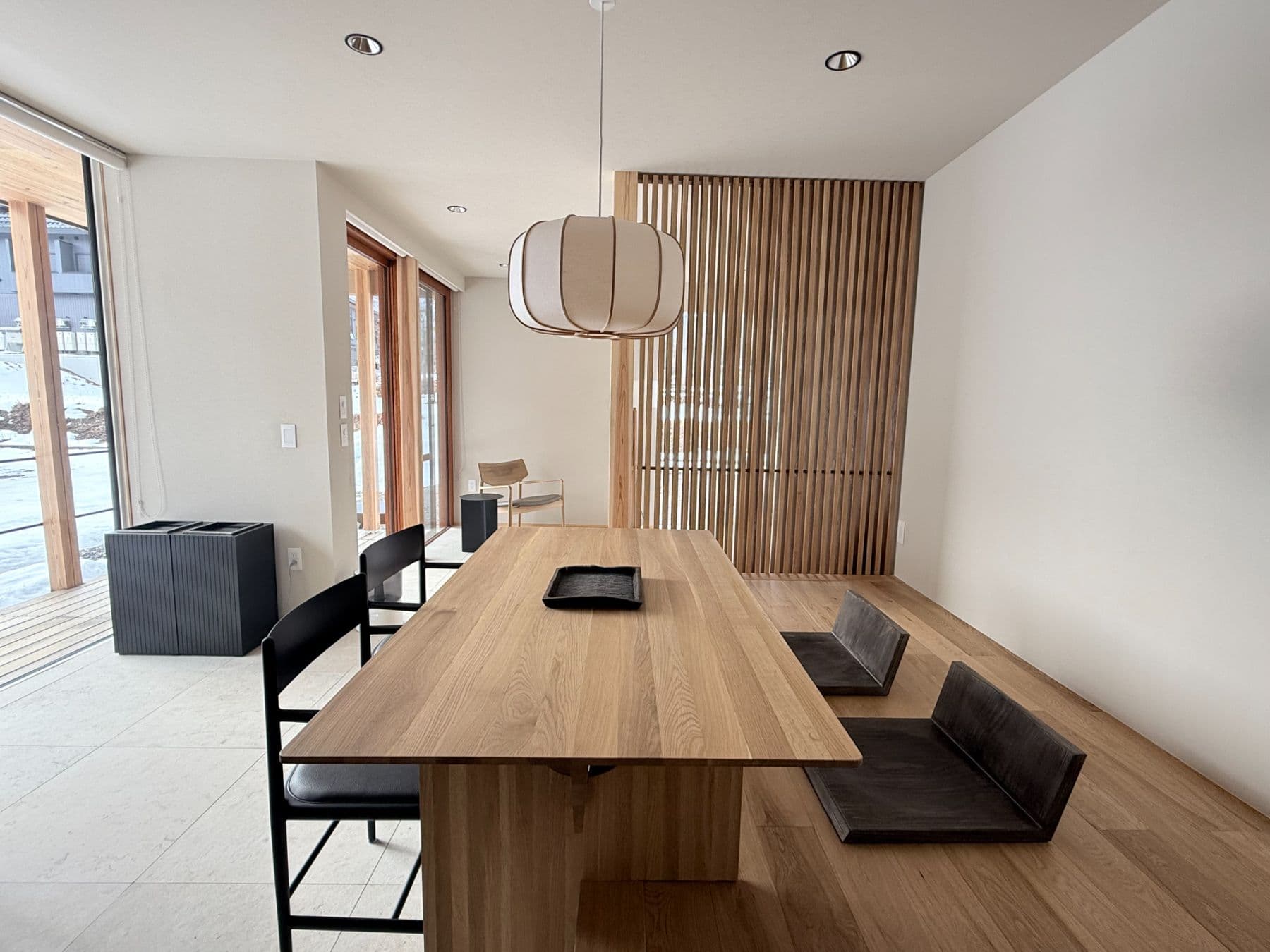 A Japanese-modern dining room with a pendant light, long wooden table, wood-slat partition and bright windows.