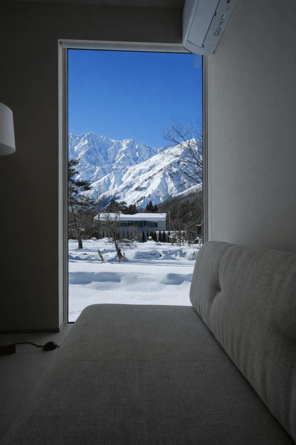 A large window overlooking the snow-capped Hakuba mountains and winter garden, with a sofa in the foreground.